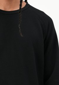 Chemise noire à manches longues en tricot gaufré avec un col rond et des manches raglan, présentant un tissu texturé et un design minimaliste.