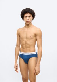 Ανδρικά εσώρουχα σε ναυτικό μπλε χρώμα με λευκή ελαστική μέση που φέρει το λογότυπο "TOMMY HILFIGER". Το ύφασμα φαίνεται λείο και εφαρμοστό. Το μοντέλο στέκεται όρθιο.