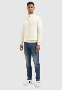 Man staat gekleed in een off-white ritsneck sweater, vervaagde blauwe spijkerbroek en witte sneakers met zwarte accenten, tegen een effen lichte achtergrond.