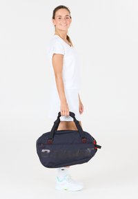 Ung kvinde i hvid sportstøj, der holder en sort Babolat tennis taske, står og smiler foran en ensfarvet hvid baggrund.