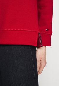 Czerwona bluza z teksturą, z bocznym rozcięciem i małą metką z logo. Połączona z ciemnym dżinsem, podkreślająca kontrastujące kolory.