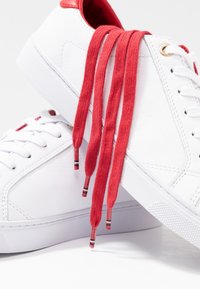 Un par de zapatillas de piel blancas con cordones rojos y forro interior rojo, una zapatilla descansando sobre la otra en una superficie blanca.