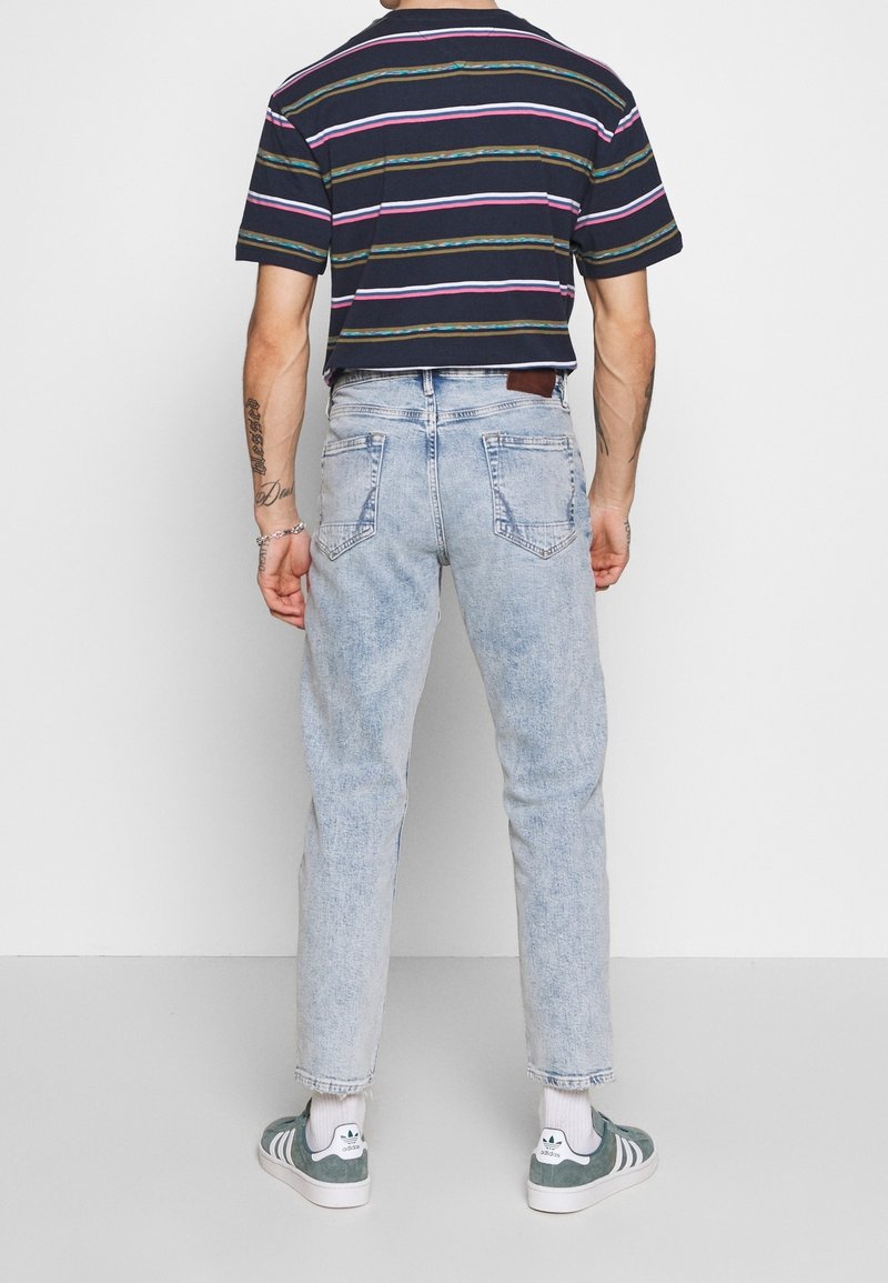 Personne portant un t-shirt rayé noir à manches courtes, un jean bleu clair, des chaussettes blanches et des baskets Adidas vertes, debout dos à la caméra.