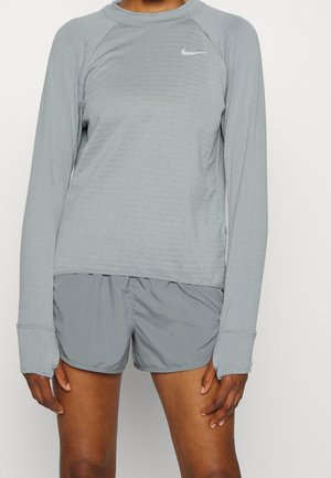 Personne portant un t-shirt Nike à manches longues gris clair avec des trous pour les pouces et un short de sport gris clair assorti, debout devant un fond uni.
