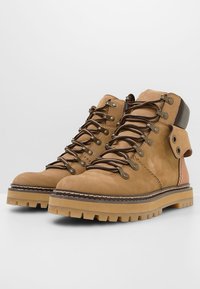 Hnědé kožené turistické boty s béžovými šňůrkami, kovovými očky a robustní podrážkou. Design zahrnuje polstrovaný límec a kontrastní detail na patě.