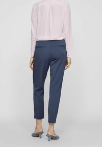 Pantalon fuselé bleu marine à texture lisse, avec deux poches arrière et une taille mi-haute, assorti à un chemisier rose clair et des talons bleus.