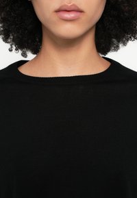 Čierny pletený sveter s okrúhlym výstrihom, ktorý má mäkkú textúru a priliehavý strih okolo goliera, zvýrazňujúci hladkú látku a jednoduchosť.