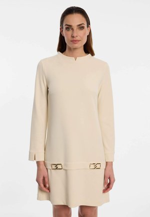 Woman wearing a beige long-sleeve dress with a small front slit neckline and gold rectangular buckle details near the hem.