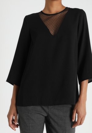 Schwarze Bluse mit langen Ärmeln und transparentem gepunktetem V-Ausschnitt, getragen zu grauen, karierten Hosen, dargestellt von den Schultern bis zur Hüfte.