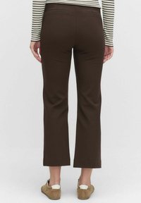 Persoon in bruine cropped broek, beige sneakers en een gestreept Shirt met lange mouwen, staande met de rug naar de camera tegen een effen achtergrond.