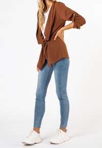 Brun, lättviktsjacka med öppen front, knytning i midjan och uppvikta ärmar. Kombinerad med ljusblå tajta jeans och vita sneakers.