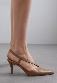 Chaussure à bout pointu beige au design épuré, avec un talon court et une bride délicate autour de la cheville. Fabriquée en matériau lisse et brillant.
