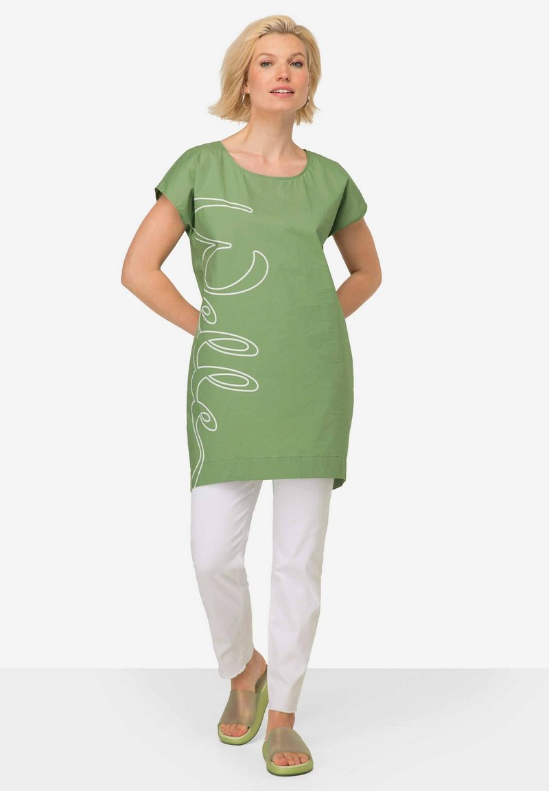 Zelená tunika s bílým písmem, krátké rukávy, volný střih, v kombinaci s bílými kalhotami a zelenými nazouváky, hladká textura látky.