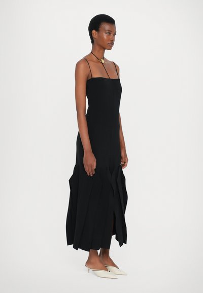 Μαύρο, αμάνικο φόρεμα με δομημένο σχέδιο που διαθέτει ασύμμετρο, φουσκωτό τελείωμα και υφασμάτινη λεπτομέρεια. Συνδυασμένο με λευκά, μυτερά παπούτσια.