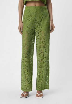 Pantalones de encaje verdes con un ajuste holgado, que presentan patrones florales y circulares, diseño de pierna ancha y una textura suave. Calzado blanco visible.