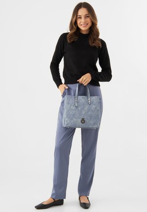 Blauwe stoffen boodschappentas met twee handvatten bovenaan, knoopaccenten en een patroonstructuur, vastgehouden door een persoon in een zwarte trui en blauwe broek.