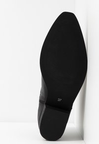 Chaussure en cuir noir avec une semelle en caoutchouc texturée, présentant un bout pointu et une étiquette de taille 37. Fini lisse, design minimaliste.