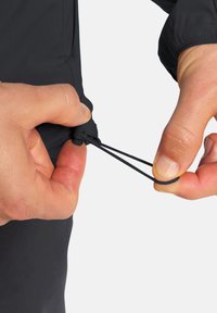 Veste noire avec un cordon élastique ajustable, montrée dans une main, présentant une texture lisse et un design épuré, mettant l'accent sur le mécanisme de serrage.