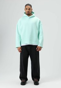 Sweat à capuche oversize vert menthe avec une poche avant, porté par-dessus une chemise blanche, accompagné d'un jean noir ample et de chaussures noires épaisses.