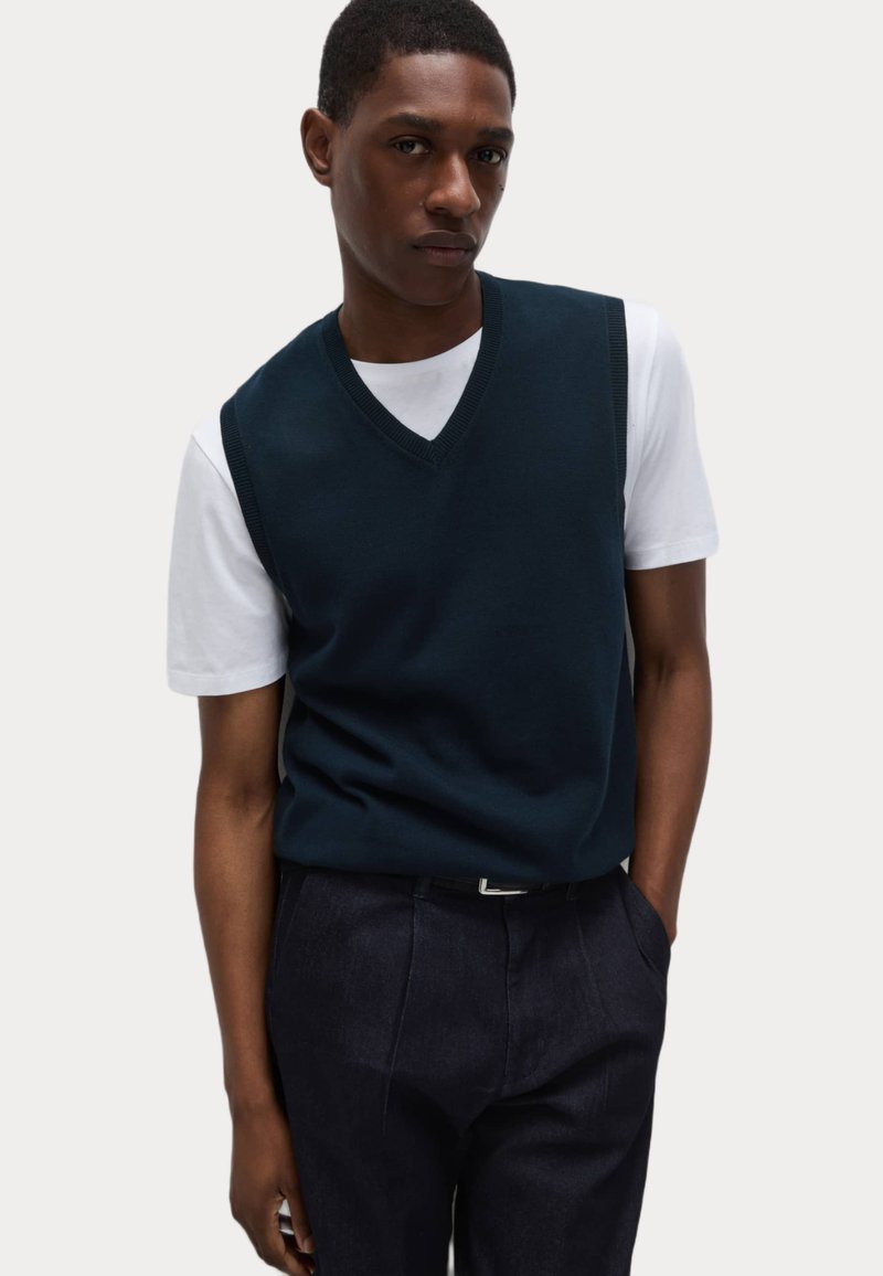 Maglione senza maniche blu navy con scollo a V, indossato sopra una camicia bianca a maniche corte, abbinato a pantaloni scuri. Tessuto a maglia texturizzata.