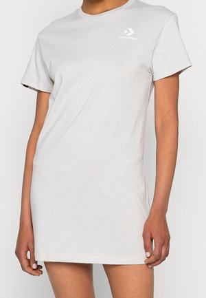 Persoon die een effen lichtgrijze korte mouw T-shirtjurk draagt met een klein wit Converse-logo op de bovenste linkerborst.