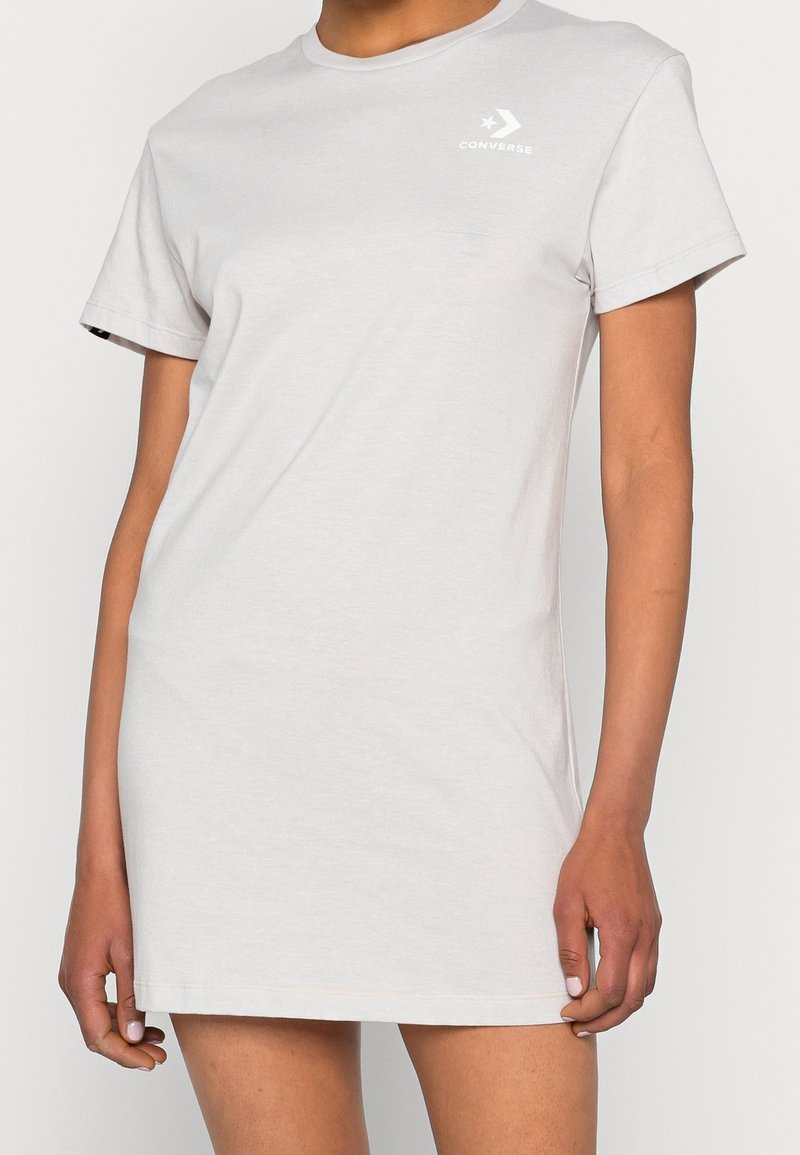 Personne portant une robe T-shirt à manches courtes gris clair unie avec un petit logo blanc Converse sur la partie supérieure gauche de la poitrine.
