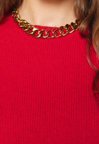 Punainen, ribbineulottu neulepaita, jossa on kullanvärinen ketjukaulus. Kangas on teksturoitu ja kauluksessa on isot, kiillotetut lenkit.