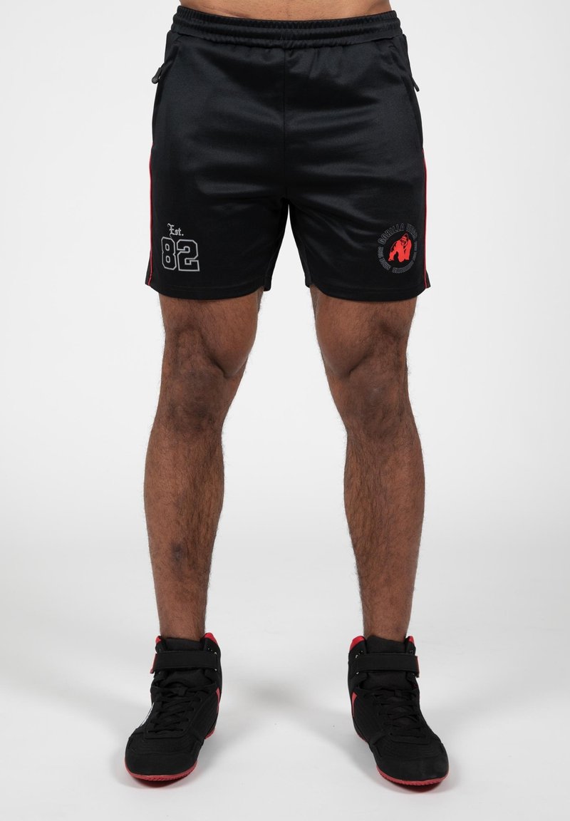 Shorts de sport noirs avec des accents rouges sur les côtés, arborant un graphique gris "Est. 82" et un logo rouge. Fabriqués en tissu lisse et léger.