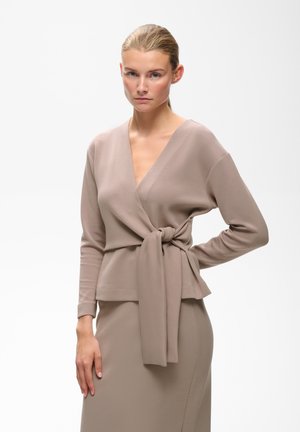 Kvinde med blondt hår i en taupe omslags-stil top bundet ved taljen og matchende nederdel står mod en hvid baggrund.