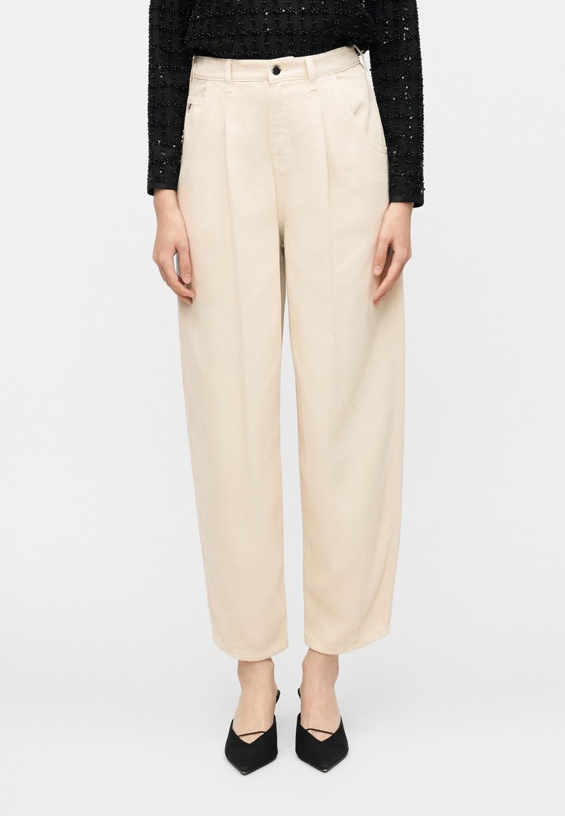 Μπεζ παντελόνι φαρδιάς γραμμής με πιέτες, κατασκευασμένο από υφή ύφασμα. Διαθέτει κούμπωμα με κουμπί και πλαϊνές τσέπες. Φορεμένο με μαύρες μυτερές γόβες.