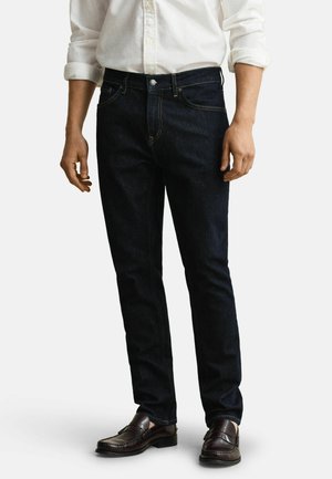 Mann trägt dunkelblaue, eng geschnittene Jeans, weißes Hemd mit hochgekrempelten Ärmeln und dunkelbraune Leder-Loafer, steht vor weißem Hintergrund.
