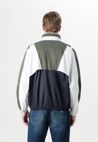Fargeblokkert vindjakke med olivengrønne, hvite og marineblå paneler, elastisk kant og høy krage. Lett og glatt tekstur.