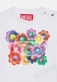 Bijela majica s živahnim cvjetnim grafikom i cvjetovima s 3D efektom u raznim bojama, te crvenom oznakom "DIESEL" na izrezu.