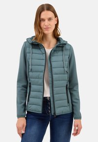 Polstret teal jakke med hætte, der har lynlås foran, sorte detaljer og elastiske manchetter. Båret over en hvid top og denim jeans.