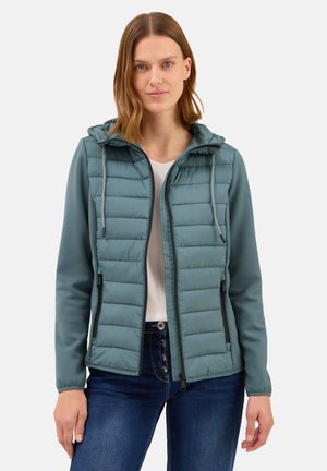 Giacca imbottita teal con cappuccio, dotata di zip frontale, accenti neri e polsini elasticizzati. Indossata sopra una maglietta bianca e jeans in denim.