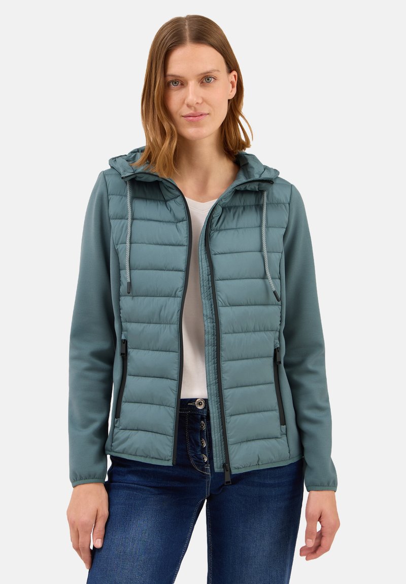 Polstret teal jakke med hætte, der har lynlås foran, sorte detaljer og elastiske manchetter. Båret over en hvid top og denim jeans.