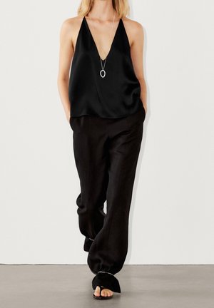 Woman in loose black halter top and wide black pants with silver anklets, walking against plain white background.