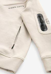 Vaaleanbeige huppari, jossa on ribbaustaskut, vetoketjullinen tasku hihassa mustilla yksityiskohdilla ja painetut koordinaatit kankaassa.