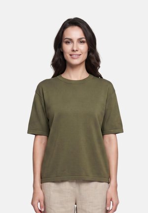 Donna con capelli scuri e mossi che indossa una camicia verde oliva a maniche corte e pantaloni beige chiaro, in piedi davanti a uno sfondo bianco.