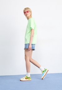 Neonově zelené oversized tričko, džínové šortky s třepícími se okraji a vícebarevné tenisky. Doplňky zahrnují růžové sluneční brýle.