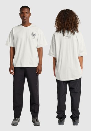 Voor- en achterkantweergave van twee modellen die witte graphic t-shirts en zwarte broeken dragen, met grijze wandelschoenen op een effen achtergrond.