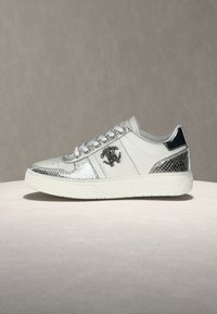 Άσπρο sneaker χαμηλού προφίλ με ασημί λεπτομέρειες σε σχέδιο φιδιού, μαύρο λογότυπο στο πλευρό, λευκά κορδόνια και παχύ λευκό πέλμα σε ουδέτερο φόντο.