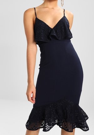 Femme portant une robe noire ajustée sans manches avec des volants en dentelle au niveau de l'encolure et de l'ourlet, debout devant un fond gris uni.