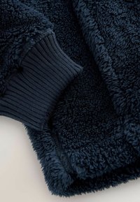 Tessuto in pile blu navy con una superficie strutturata e soffice. Il polsino presenta un design liscio a coste per una vestibilità aderente.