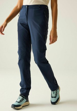 Osoba ubrana w granatowe spodnie outdoorowe i jasnoniebieskie buty trekkingowe idzie po jednolitym jasnym tle.