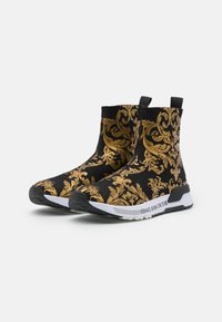 Svarta ankelboots med intrikata guldfärgade blomsterornament, elastisk överdel och en vit sula med "VERSACE JEANS COUTURE"-märke.