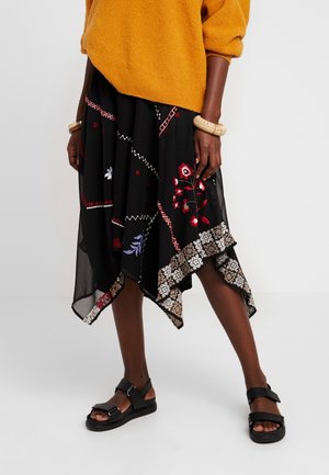 Person iført en sennepsgul sweater, en sort asymmetrisk nederdel med blomster- og geometriske mønstre, sorte sandaler og en beige armring.