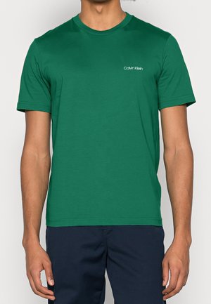T-shirt en coton vert avec col rond et manches courtes, arborant un logo Calvin Klein blanc sur le côté gauche de la poitrine. Texture lisse.