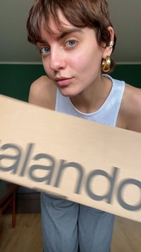 Caja de cartón marrón que muestra "Zalando" en letras gris oscuro. La persona la sostiene mientras lleva puestos pantalones gris claro y una camiseta sin mangas blanca.