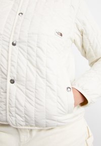 Veste matelassée blanche avec boutons-pression métalliques, une poche poitrine visible avec le logo The North Face et une main dans la poche latérale.